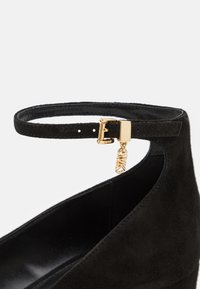 Zapato de tacón alto de ante negro con una delgada correa de tobillo y detalle de hebilla de metal dorado y charm. Superficie texturada con un interior suave.