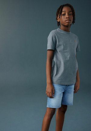 Băiat cu păr împletit, purtând un tricou gri cu mâneci scurte și pantaloni scurți din denim albastru deschis, stând în fața unui fundal simplu de culoare teal închis.