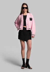 Cardigan rosa a coste con accenti neri e ricamo della 'W', indossato sopra un crop top nero e una gonna nera asimmetrica, con sandali con tacco alto.