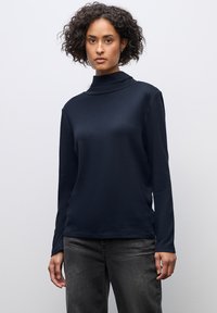 Marineblaues Langarm-Turtleneck-Oberteil aus weichem Material, mit einem Stehkragen und einer lässigen Passform, kombiniert mit dunkelgrauen Jeans.