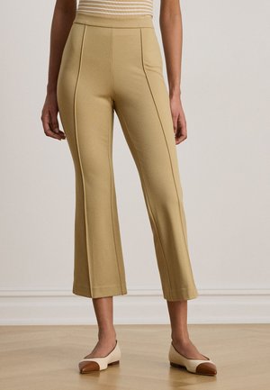 Kvinne iført beige korte bukser med frontsøm og beige flate sko med brune tåhetter som står på tregulv.