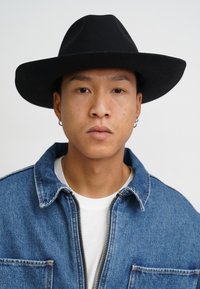 Schwarzer, breitkrempiger Filzhut, Jeansjacke mit silberner Reißverschluss, darunter ein weißes Hemd, getragen vor einem schlichten, hellen Hintergrund.