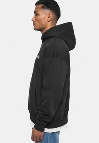 Schwarzer Hoodie aus Baumwolle, mit Kängurutasche, gerippten Bündchen und Kapuze mit Kordelzug. Von der Seite gezeigt.