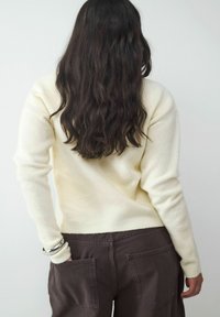 Helle, gelbe Strickjacke mit langen Ärmeln, getragen mit dunkelbraunen Hosen. Sie hat eine einfache, glatte Textur und einen lässigen Schnitt.