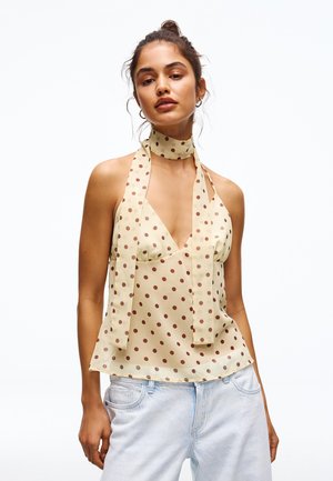 Junge Frau mit hochgestecktem Haar, die ein ärmelloses beigefarbenes Neckholder-Oberteil mit braunen Punkten und hellblaue Jeans vor weißem Hintergrund trägt.