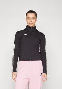 Črna zip-up jakna z visokim ovratnikom, ki ima beli Adidas logotip in tri bele črte vzdolž rokavov. Gladek material, prilegajoča zasnova.