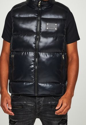 Mann trägt eine schwarze, glänzende, gepolsterte, ärmellose Weste, die zugezippt ist, über einem schwarzen Kurzarmhemd und abgenutzten schwarzen Jeans mit Reißverschlusstaschen.