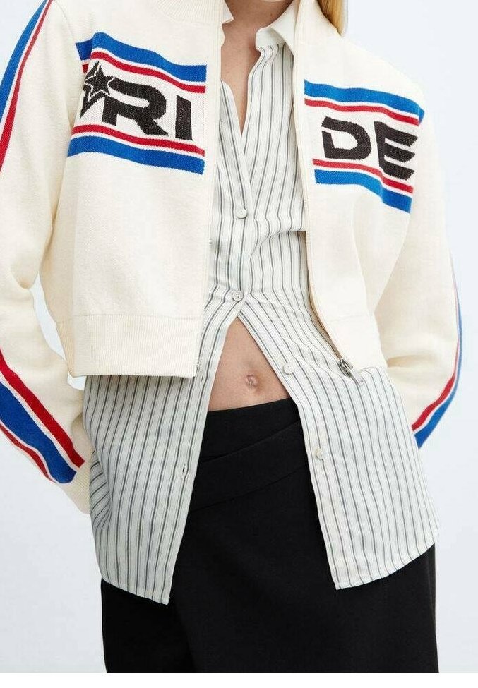 Bílá pletená kardiganka s červenými a modrými pruhy, se slovem "RIDE" v černé barvě, nošená přes pruhovanou košili s knoflíky, spárovaná s černými kalhotami.