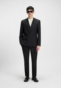 Schwarze Doppeltbreasted-Anzugjacke mit passender Hose, begleitet von einem weißen Hemd. Bemerkenswerte Merkmale sind flache Taschen und elegante schwarze Schuhe.