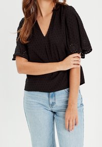 Blouse noire avec un décolleté en V, des manches courtes évasées avec des accents brodés, et un motif en relief à pois, associée à un jean bleu clair.