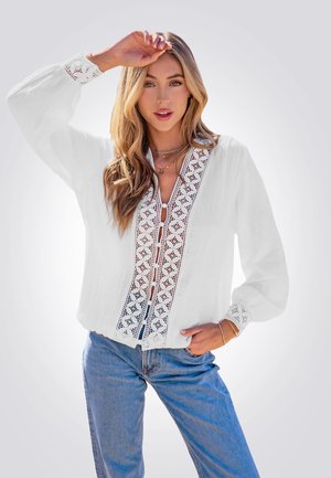 Blusa blanca con un canesú de encaje detallado, cierre de botones y mangas largas. Presenta una tela texturizada y un corte relajado. Combinada con jeans azules.