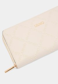 Cream wallet with textured surface featuring a repeated embossed logo. Gold-tone zipper and hardware. Slim rectangular shape.