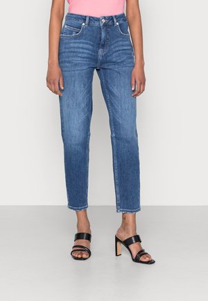 Jeans de mezclilla azul claro con corte recto, cinco bolsillos y dobladillos deshilachados. Combinados con sandalias negras de tacón alto y tiras.