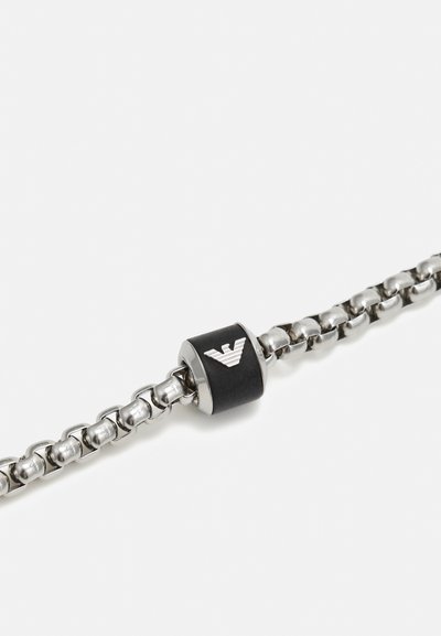 Sølvkjedearmbånd med runde lenker og en sentral svart luke med et metallisk logo. Glatt tekstur med polert finish.