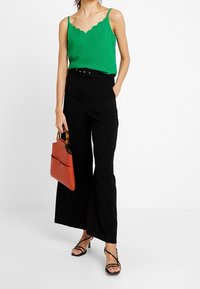 Camisola verde con bordo a scallop abbinata a pantaloni neri a vita alta con gamba larga. Gli accessori includono una borsa rossa e sandali neri con strappi.