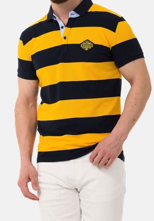 Homme portant un polo rayé horizontal jaune et noir avec un logo sur la poitrine et un pantalon blanc, la main reposée près de la poche.