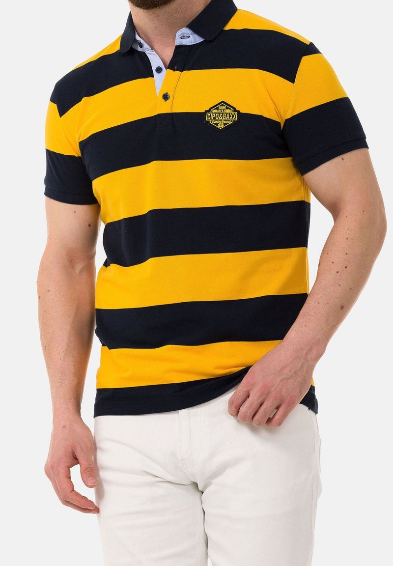 Homme portant un polo rayé horizontal jaune et noir avec un logo sur la poitrine et un pantalon blanc, la main reposée près de la poche.