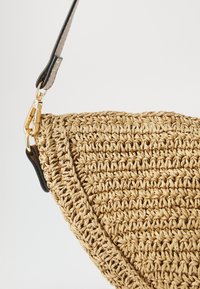 Tkaná slaměná kabelka v přirozené béžové barvě s zakřiveným designem. Disponuje černým koženým popruhem a kovovými doplňky ve zlatém tónu pro upevnění.