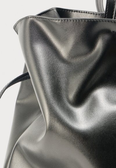 Grand sac cabas en cuir noir brillant avec plis visibles et texture lisse, présentant des coutures sur le bord supérieur et des poignées fines.