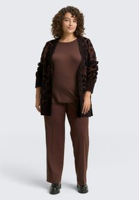 Brauner Cardigan mit Leopardenmuster über einem braunen Oberteil und passenden Hosen mit weitem Bein. Glatte Textur mit Knöpfen am Cardigan und klobigen schwarzen Stiefeln.