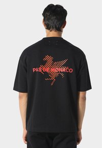 Man in een zwart oversized T-shirt met een rood gestreepte gevleugelde paard grafiek en de tekst "PAS DE MONACO" op de achterkant, staand tegen een grijze achtergrond.