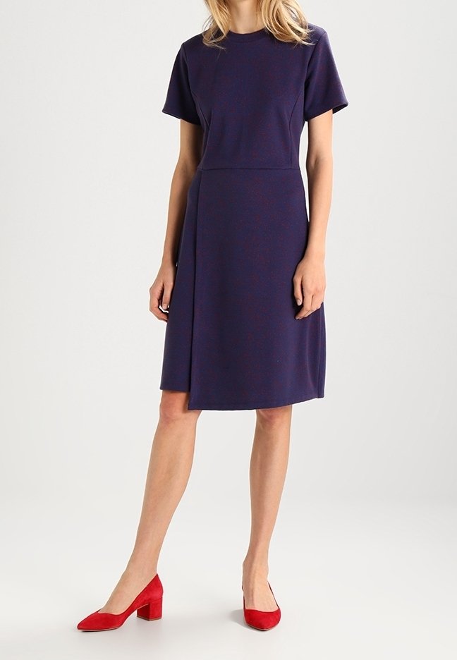 Femme portant une robe bleu marine courte arrivant au genou avec manches courtes, associée à des chaussures rouges à talons blocs, debout devant un fond clair uni.