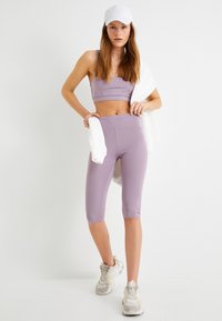 Lavendelcroppad sport-bh och högmidjade cykelshorts, tillverkade av stretchigt material. Modellen håller en vit sweatshirt och bär en vit keps.