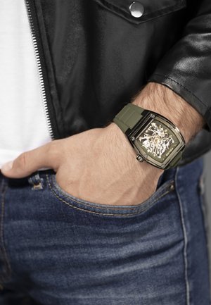 Orologio con cinturino in silicone verde e quadrante metallico quadrato, con un design scheletrico. Indossato al polso, abbinato a jeans e una giacca nera.