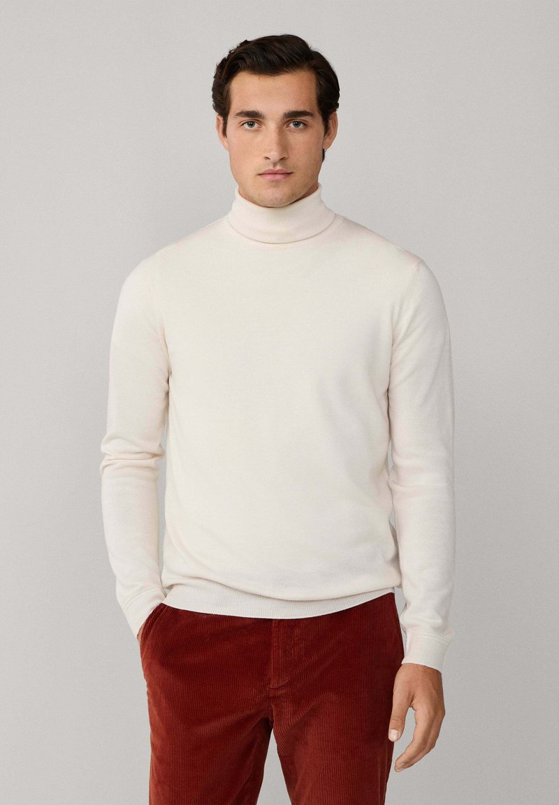 Hellbeige Rollkragenpullover aus weichem Strickstoff. Hat lange Ärmel und eine figurbetonte Passform. Getragen mit roten Cordhosen.