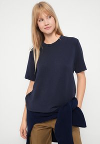 Ξανθιά γυναίκα που φοράει μια σκούρα μπλε κοντομάνικη μπλούζα με ένα σκούρο μπλε πουλόβερ δεμένο στη μέση και καφέ παντελόνια, κοιτάζοντας στο πλάι.