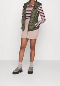 Kvinna som bär en olivgrön pufferväst över en randig långärmad tröja, beige texturerad minikjol, beige strumpor och olivgröna loafers.