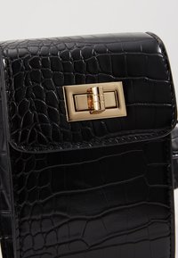 Svart handväska i krokodilprägling med en glänsande yta. Har ett guldtonat rektangulärt lås och en strukturerad design. Texturerad yta.