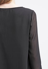 Czarna, lekka bluzka o gładkiej, przezroczystej fakturze i skromnym dekolcie. Tył ma prosty, czysty krój.