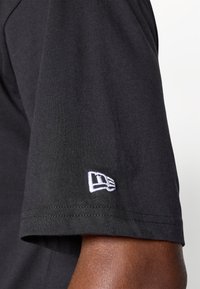 Svart kortermet t-skjorte med en liten hvit brodert logo på ermet; laget av bomull med en glatt tekstur.