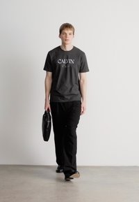 Must värvi graafiline T-särk, millel on "CALVIN" logo, kombineerituna mustade pükstega. Isik kannab musta kotti ja seisab heledal taustal.