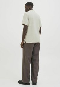 Beige polo skjorte med korte ærmer, ribbet krave, sideslids, parret med løstsiddende mørkebrune bukser og sorte sko.