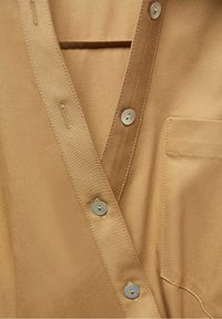 Gros plan d'une chemise marron clair à boutons, montrant les boutons, les boutonnières, le bord du col et la poche poitrine avec coutures visibles.