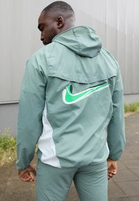 Kerge roheline tuulejakk kapuutsiga, mille seljal on suur valge ja roheline Nike logo. Jakil on sile tekstuur ja külgpaneelid.