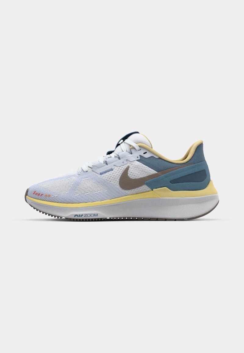 Zapatilla atlética blanca con una parte superior de malla texturada, acentos en azul claro y amarillo, y un logo de swoosh de Nike en marrón. Cuenta con una suela acolchada.
