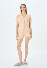 Ljusrosa kort pyjamasuppsättning med en krag skjorta och shorts. Har en texturerad tyg och en broderad "Lycka till"-grafik. Enkel design.