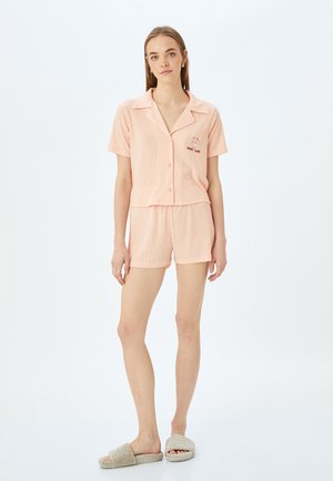 Lys pink kort pyjamasæt med en krave skjorte og shorts. Har et tekstureret stof og en broderet "Good Luck" grafik. Enkelt design.