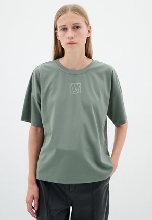Khaki vihreä lyhythihainen t-paita, jossa on pyöreä kaula-aukko ja rinnassa keskellä valkoinen geometrinen logo. Silkkinen kangas, rento malli.