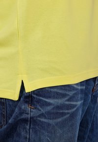 Sárga pamut póló, texturált felülettel, osztott derékkal és látható varrási részletekkel, elmosódott kék farmerral párosítva.