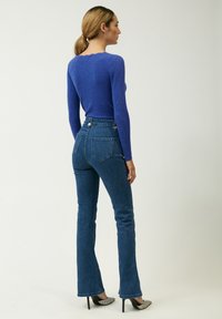 Top lungo blu a coste con collo rotondo, abbinato a jeans flare scuri a vita alta, dotati di tasche posteriori e dettagli con bottoni.