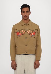 Hnědá zkrácená bunda s květinovou výšivkou v červené a modré, s špičatým límečkem a kovovými knoflíky. Texturovaná tkanina s kontrastními detaily.