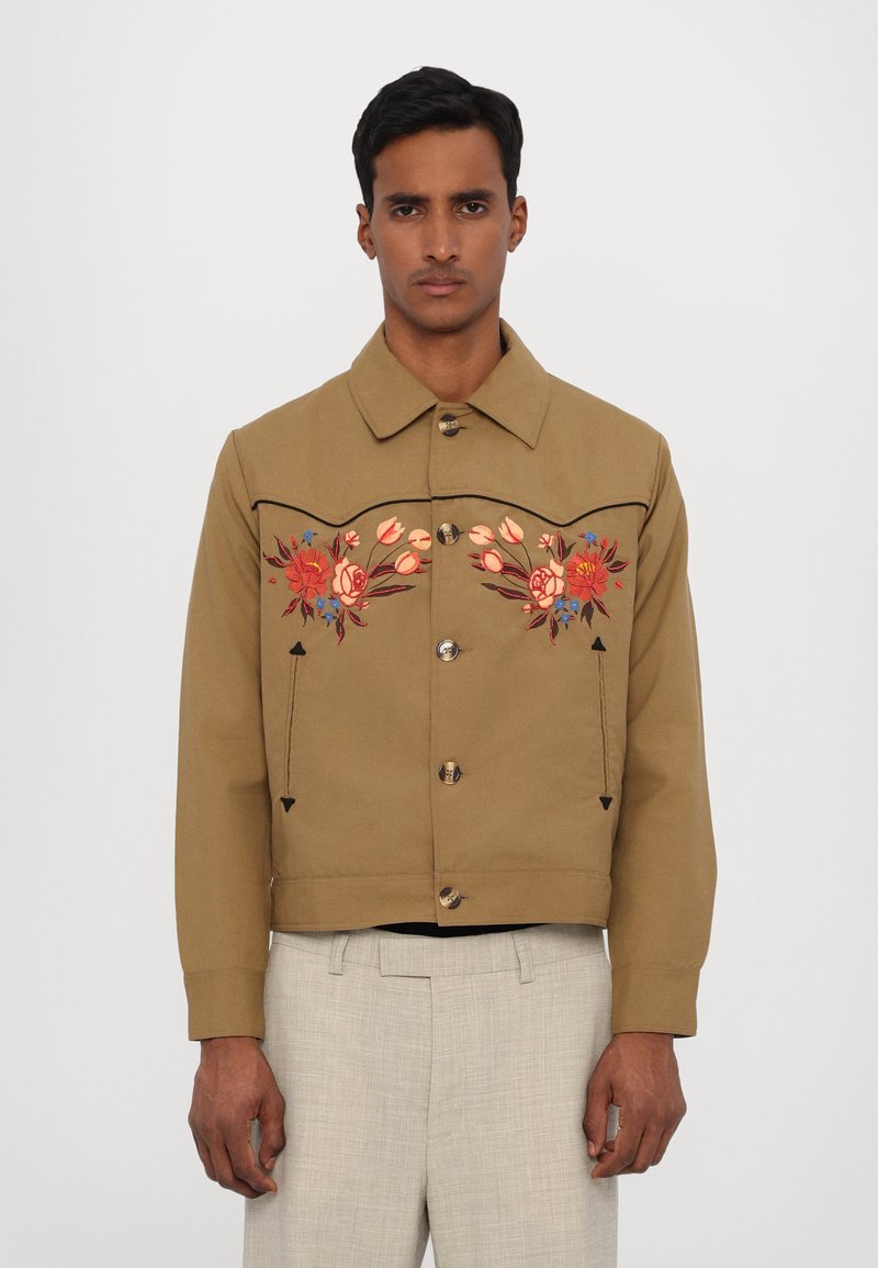 Brown cropped jacket with floral embroidery in red and blue, featuring a pointed collar and metal buttons. Textured fabric with contrasting details.