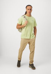 Tricou din bumbac verde deschis, pantaloni cargo bej cu buzunare, pantofi de drumeție gri și negri și un rucsac ușor. Design simplu și practic.