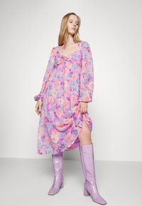 Vestido maxi floral com mangas bufantes compridas, em tons de roxo e rosa, pregas no peito, fenda lateral, acompanhado de botas lilás brilhantes até ao joelho.