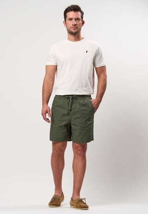 Mand stående med den ene hånd i lommen iført hvid kortærmet skjorte, olivengrønne shorts og brune bådsko på hvid baggrund.