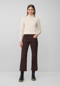 Beżowy sweter z golfem w połączeniu z ciemno-brązowymi spodniami w stylu dzwonów. Spodnie mają kieszenie z przodu i surowe wykończenie. Modelka ma na sobie czarne botki za kostkę.